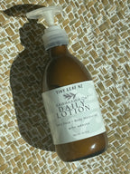 A bottle of Tiny Leaf NZ Kawakawa Oat Daily Lotion with a white pump dispenser, resting on a woven straw mat under direct sunlight.