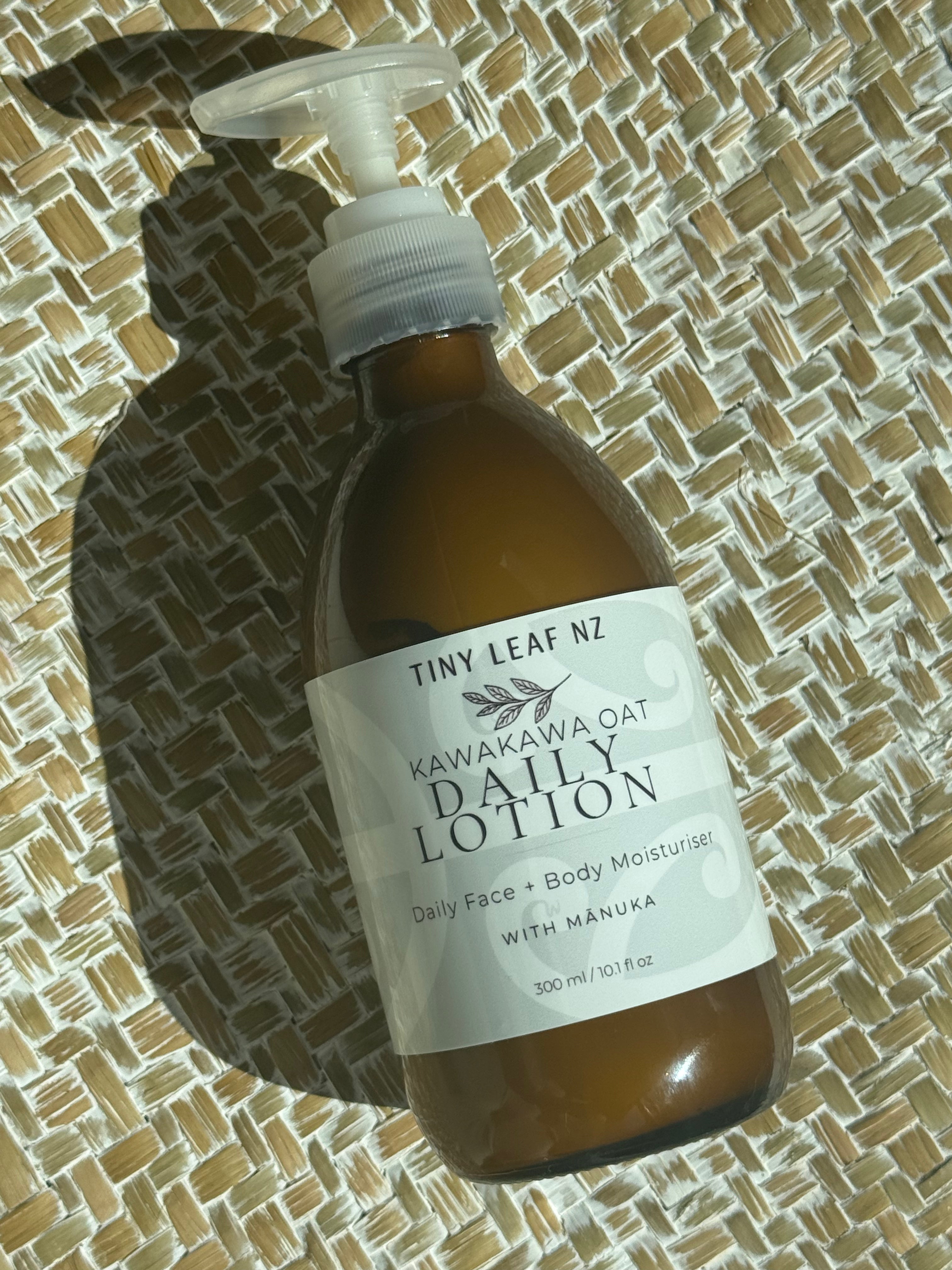 A bottle of Tiny Leaf NZ Kawakawa Oat Daily Lotion with a white pump dispenser, resting on a woven straw mat under direct sunlight.