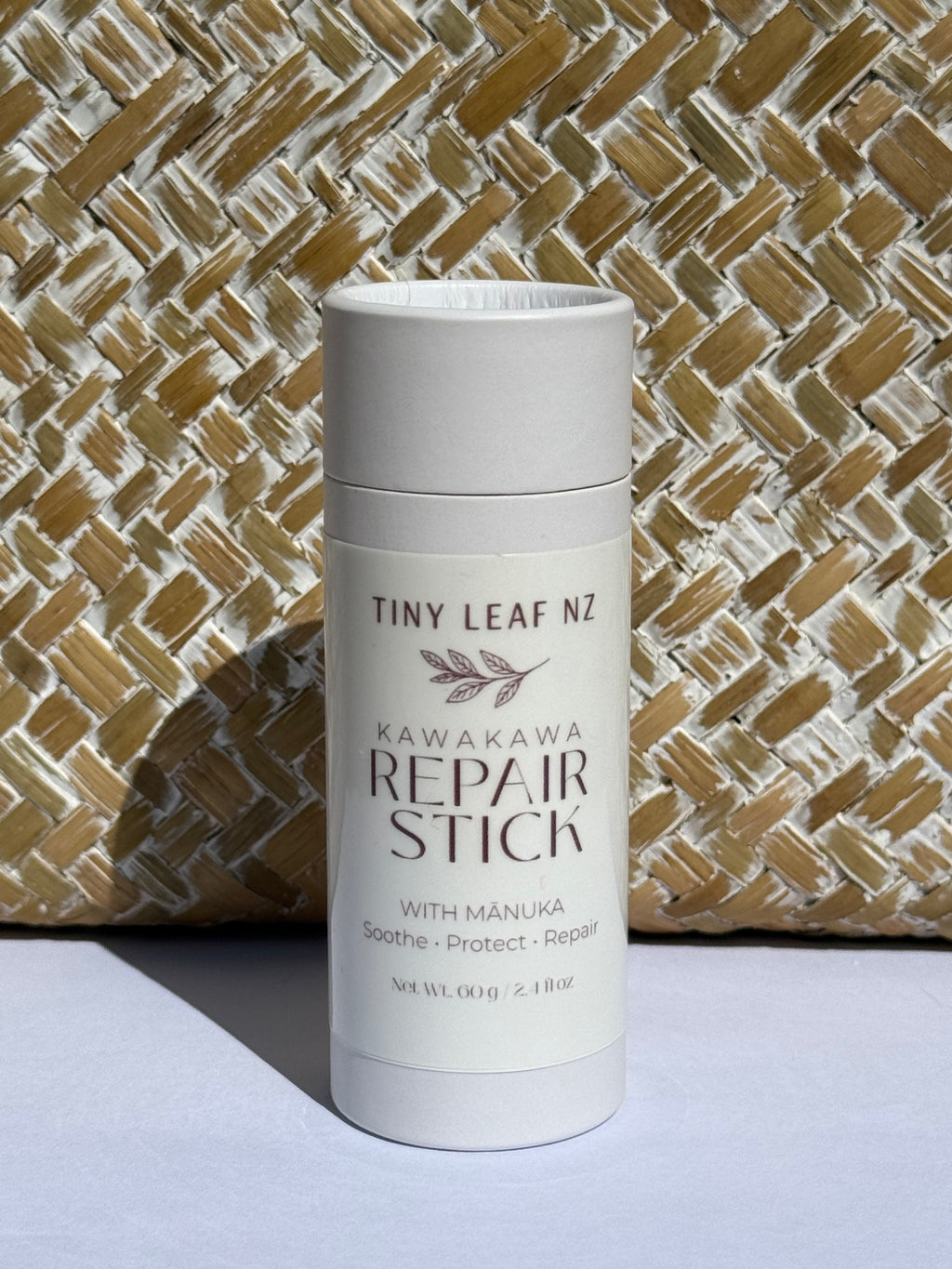 A white cylindrical deodorant-style container labeled 'Tiny Leaf Nz Kawakawa Repair Stick' stands upright against a woven straw-textured backdrop on a light surface.