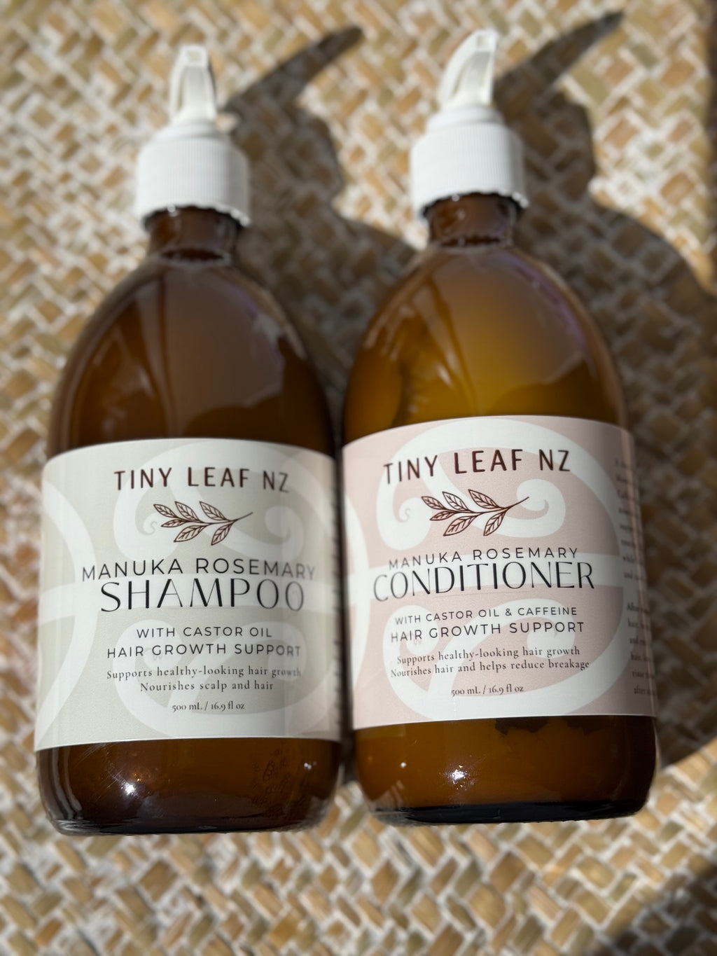 Two amber plastic bottles of Tiny Leaf NZ hair care products, Manuka Rosemary Shampoo and Manuka Rosemary Conditioner, with white caps and cream-colored labels, set against a textured beige woven background.