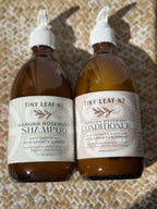 Two amber plastic bottles of Tiny Leaf NZ hair care products, Manuka Rosemary Shampoo and Manuka Rosemary Conditioner, with white caps and cream-colored labels, set against a textured beige woven background.