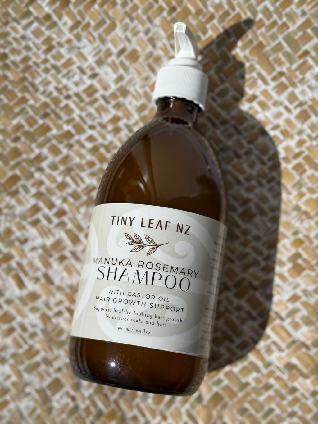 A bottle of Tiny Leaf Nz Manuka Rosemary Shampoo with a white pump dispenser, set on a textured beige and white woven mat under direct sunlight.