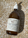 A bottle of Tiny Leaf Nz Manuka Rosemary Shampoo with a white pump dispenser, set on a textured beige and white woven mat under direct sunlight.