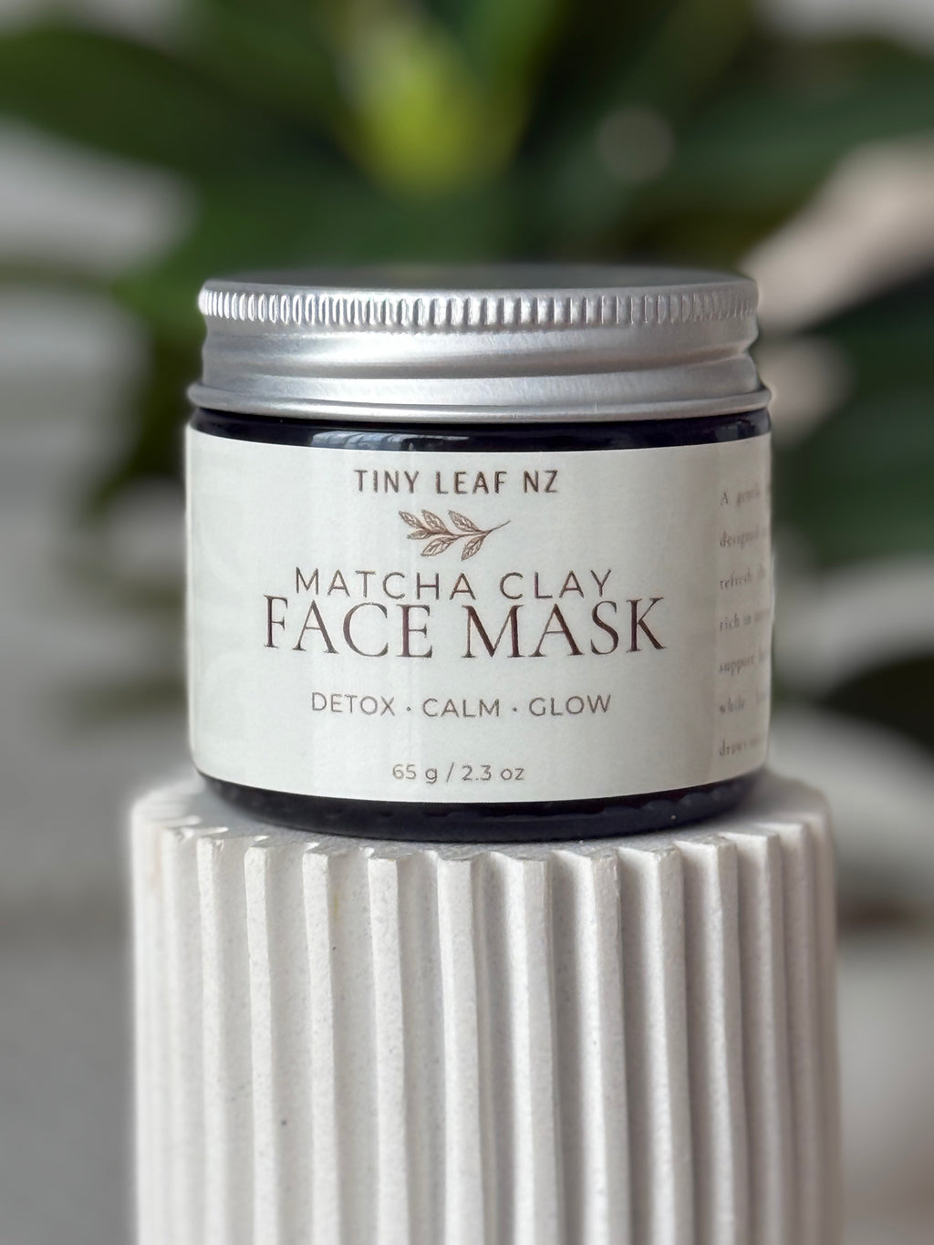 A jar of Tiny Leaf NZ Matcha Clay Face Mask sits on a ribbed white ceramic pedestal. The product is in a clear jar with a silver metal lid and a white label featuring dark brown text.