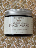 A jar of Tiny Leaf NZ Matcha Clay Face Mask resting on a woven straw mat background. The product is in a dark glass jar with a silver metal lid and a cream-colored label featuring brown text.