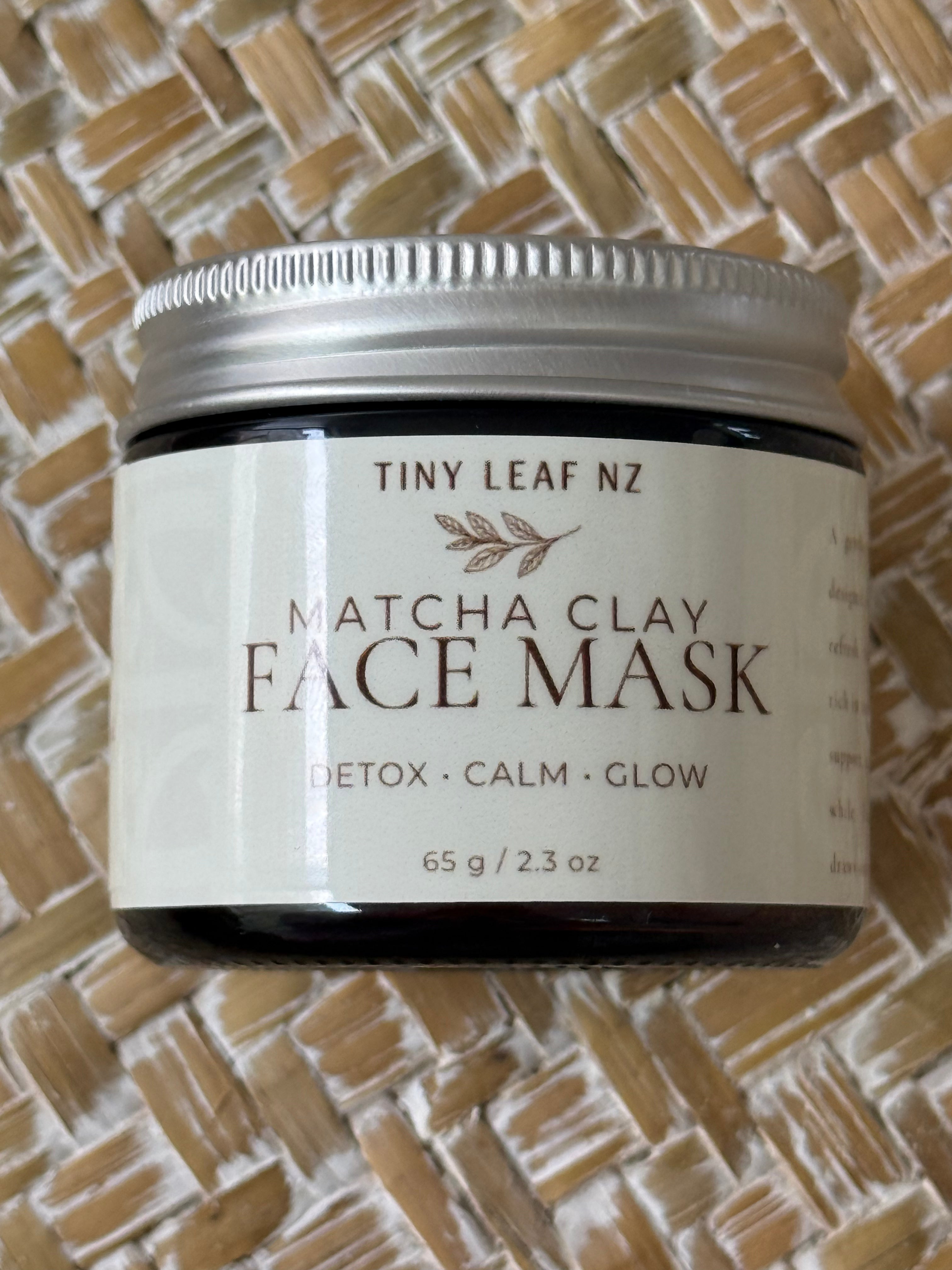 A jar of Tiny Leaf NZ Matcha Clay Face Mask resting on a woven straw mat background. The product is in a dark glass jar with a silver metal lid and a cream-colored label featuring brown text.