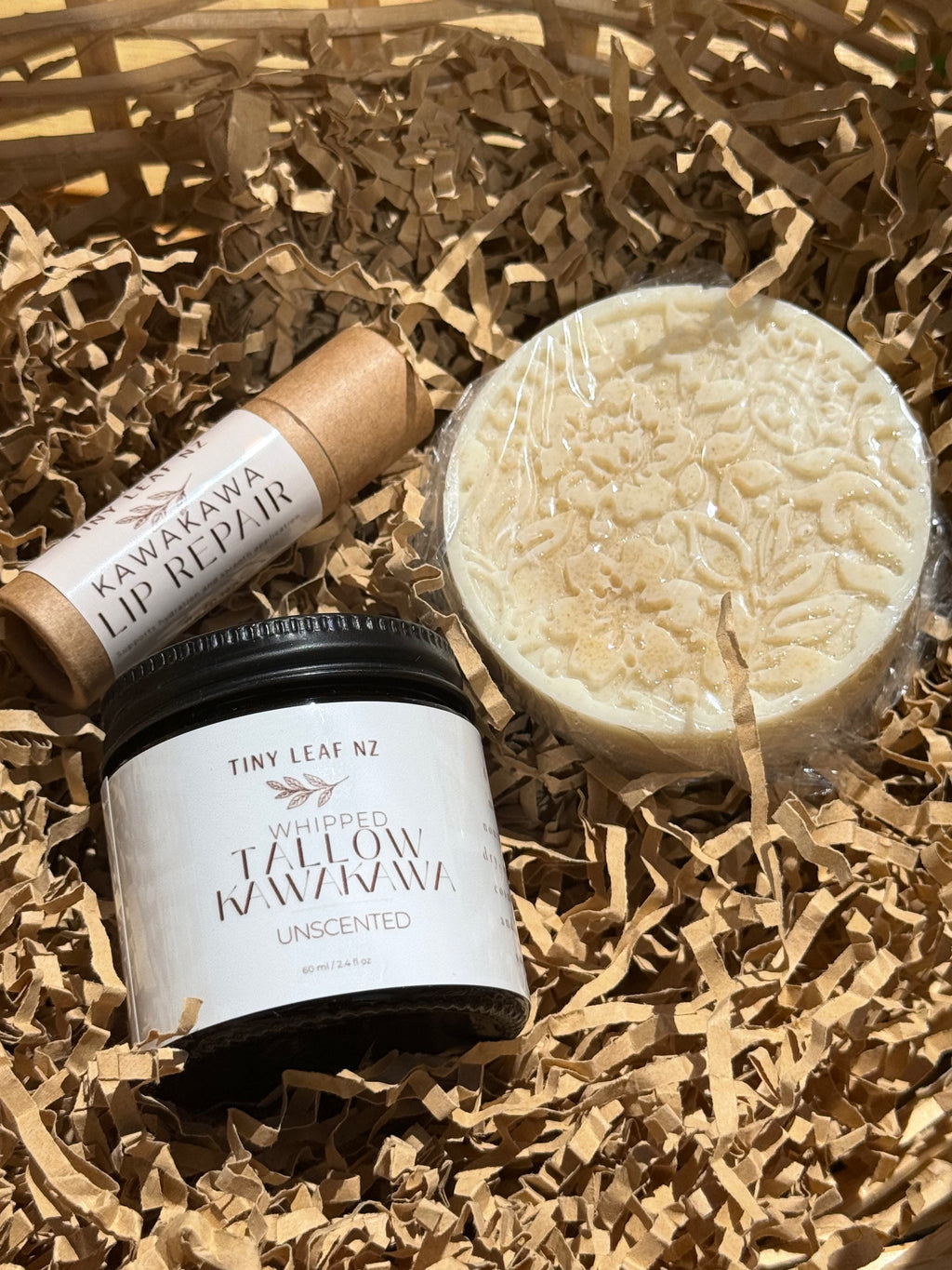 Three natural skincare products arranged in brown shredded paper: a round beige soap bar wrapped in clear plastic, a small kraft tube labeled Kawakawa Lip Repair, and a black jar labeled Tiny Leaf Nz Whipped Yellow Wax Wax Unscented.