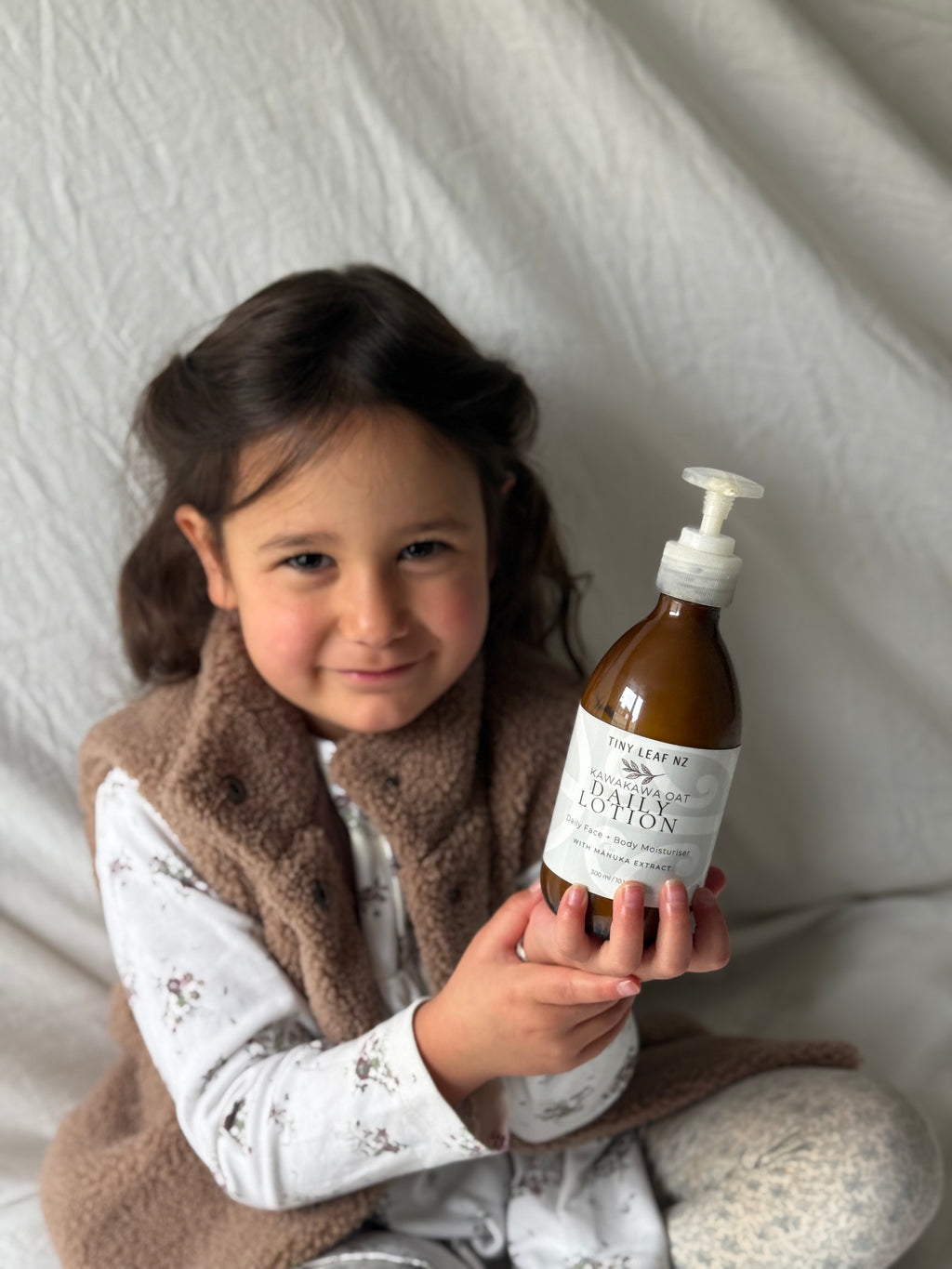 A young girl with dark hair sits against a white fabric backdrop, holding a brown pump bottle labeled 'tiny leaf face & body Lotion'. She is wearing a brown fleece vest over a white patterned long-sleeve shirt and patterned pants.