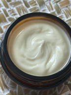 A close-up top-down view of a cream-colored cosmetic or skincare product inside an open amber glass jar, set on a woven beige and white textured surface.