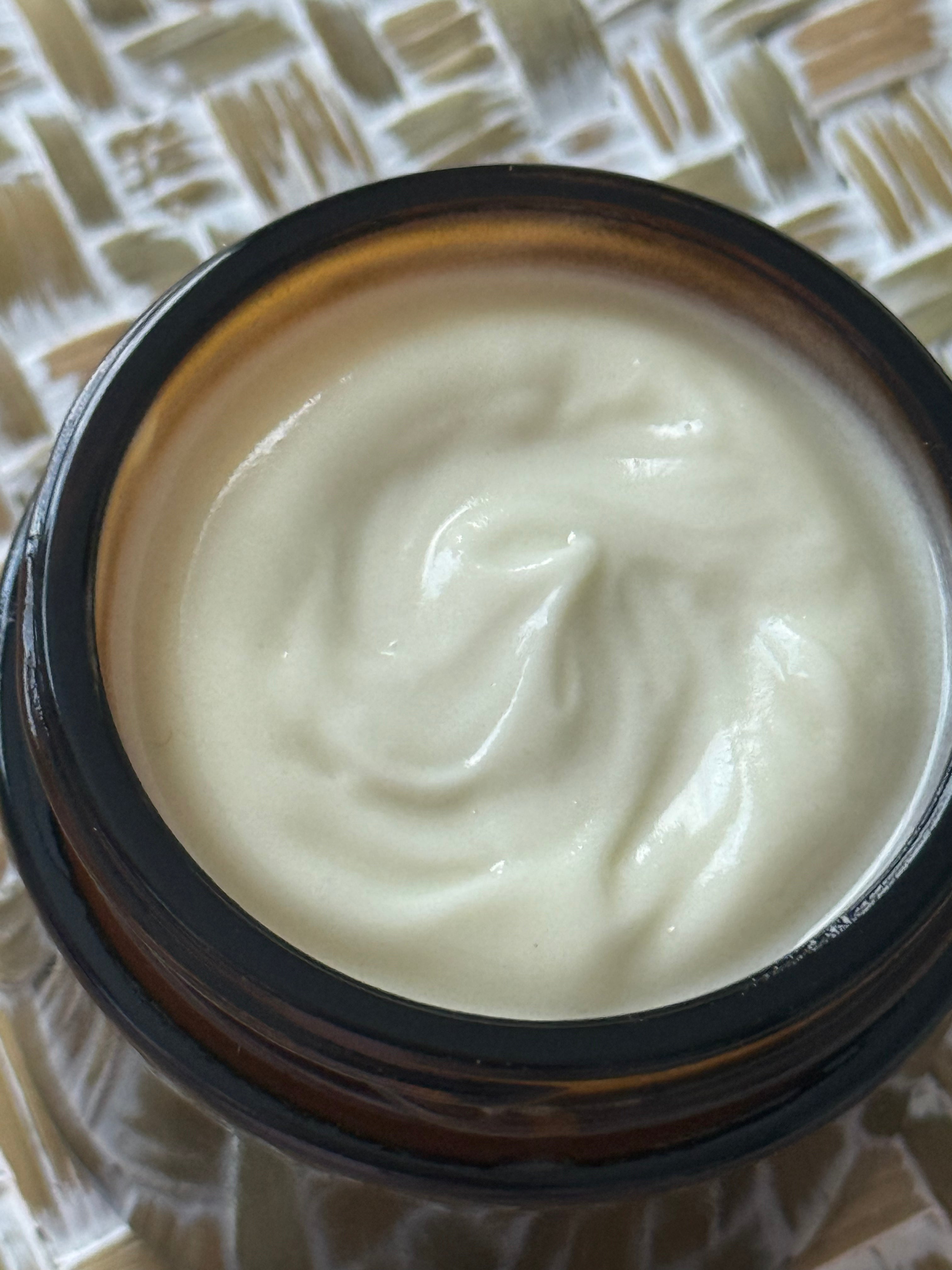 A close-up top-down view of a cream-colored cosmetic or skincare product inside an open amber glass jar, set on a woven beige and white textured surface.