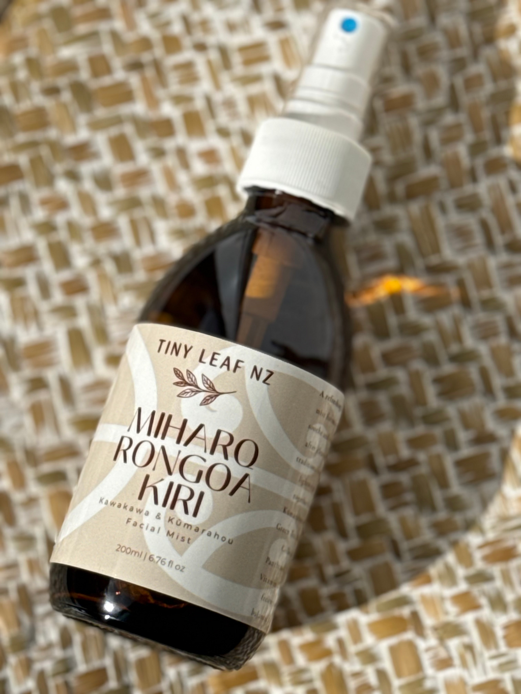 A bottle of Miharo Rongoa Kuri facial mist by Tiny Leaf NZ, featuring an amber glass bottle, white spray nozzle, and a beige label, resting on a woven brown and beige textured mat.