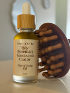 A bottle of Tiny Leaf NZ Rosemary Kawakawa Castor Hair & Scalp Oil stands on a light surface next to a wooden scalp massager.