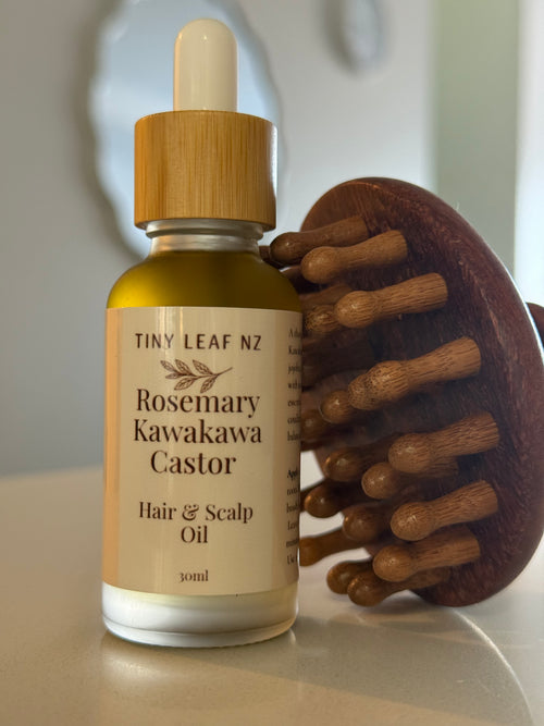 A bottle of Tiny Leaf NZ Rosemary Kawakawa Castor Hair & Scalp Oil stands on a light surface next to a wooden scalp massager.