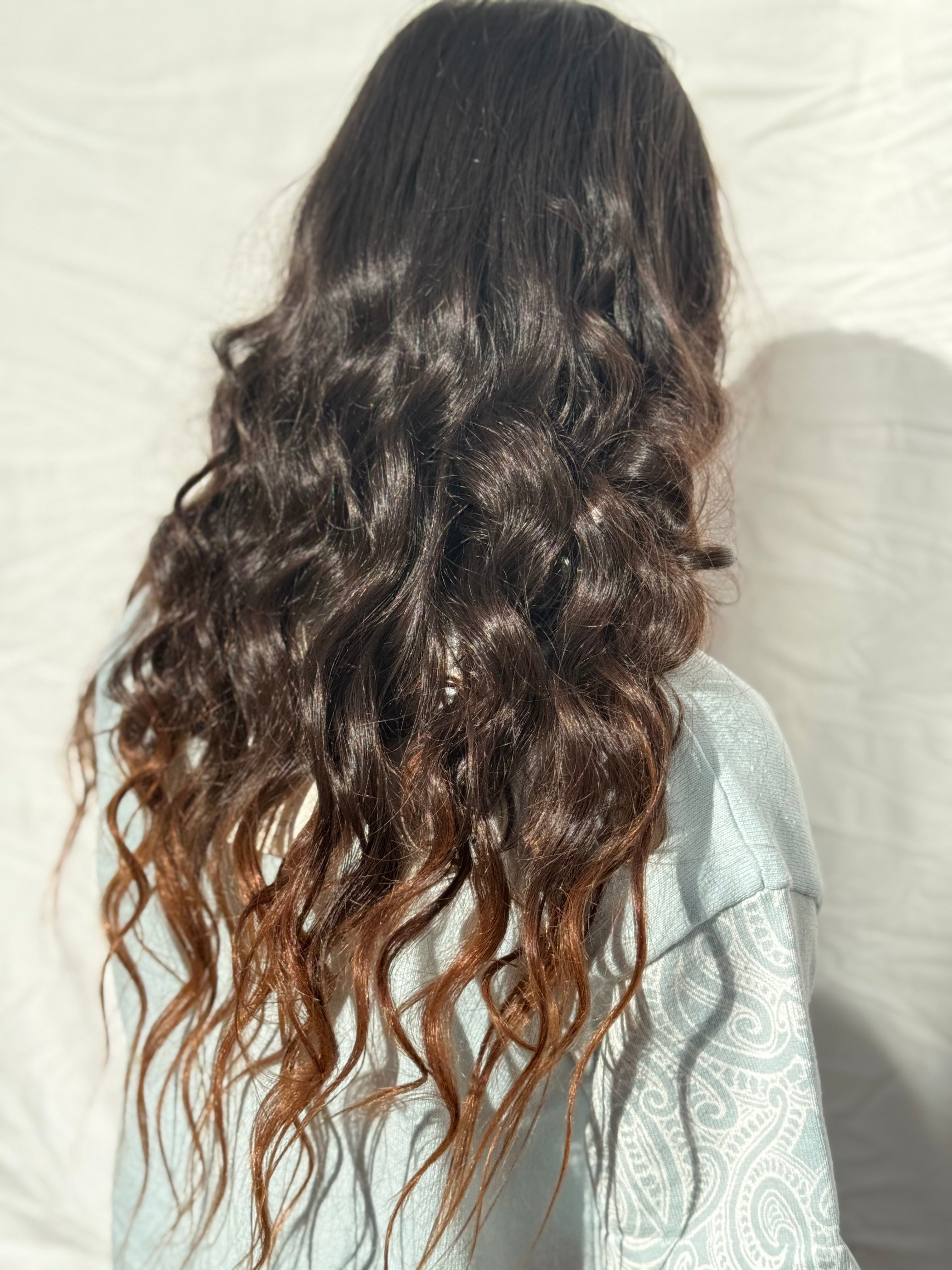 Long, wavy dark brown hair with warm caramel-brown highlights, shown from the back on a person wearing a light blue paisley top against a white fabric backdrop.