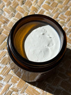 An open amber glass jar filled with a thick white cream or balm, photographed from above on a woven straw mat under direct sunlight.
