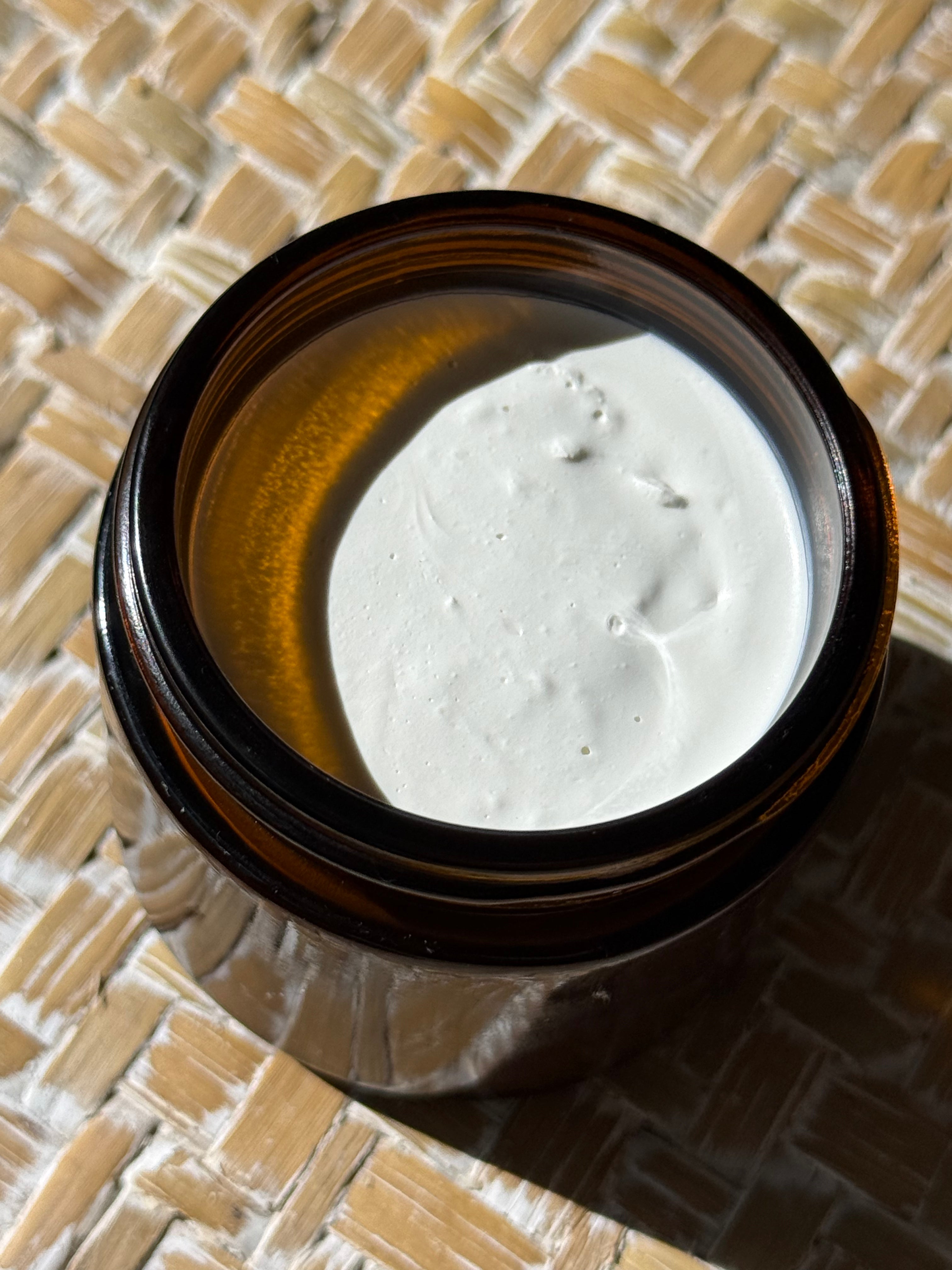 An open amber glass jar filled with a thick white cream or balm, photographed from above on a woven straw mat under direct sunlight.