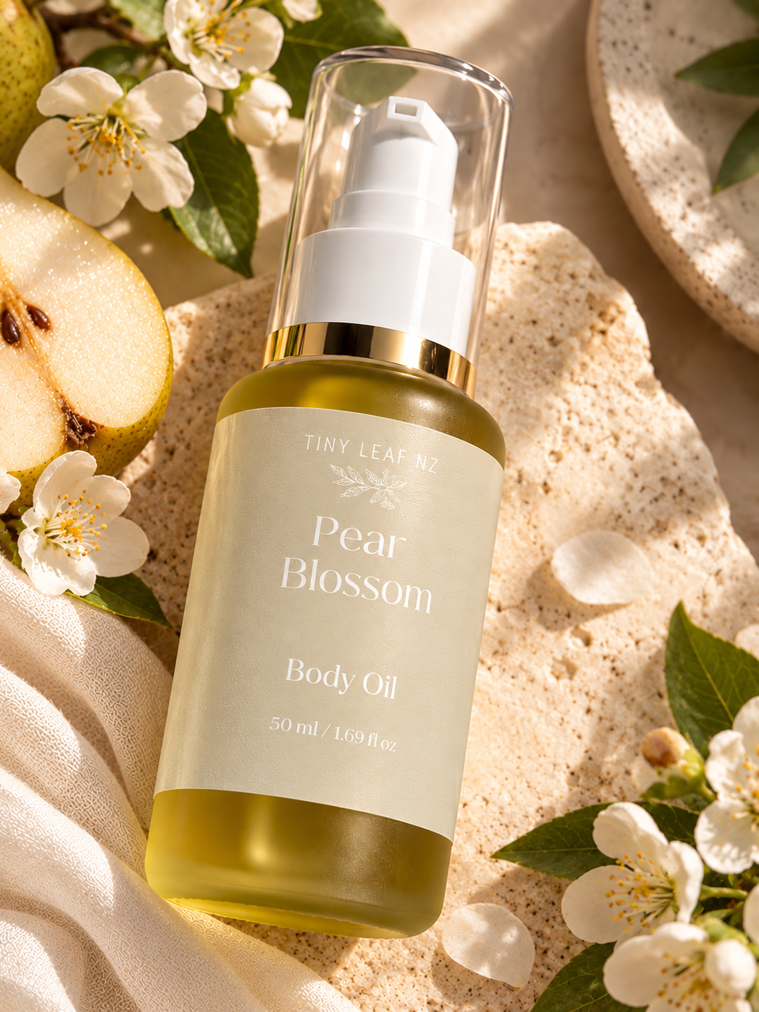 A bottle of Tiny Leaf NZ Pear Blossom Body Oil with a clear cap and white pump, filled with pale yellow-green oil, styled on a textured beige surface with white blossoms, green leaves, and a sliced pear.