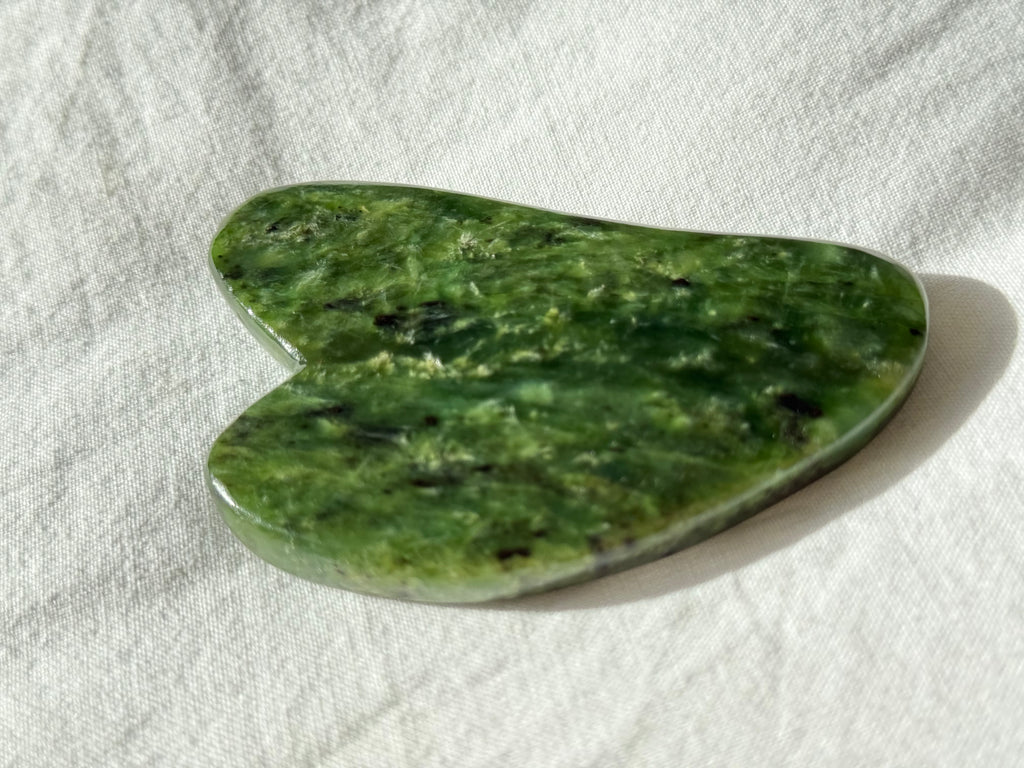 A heart-shaped green jade Gua sha massage tool resting on a textured white linen fabric background.