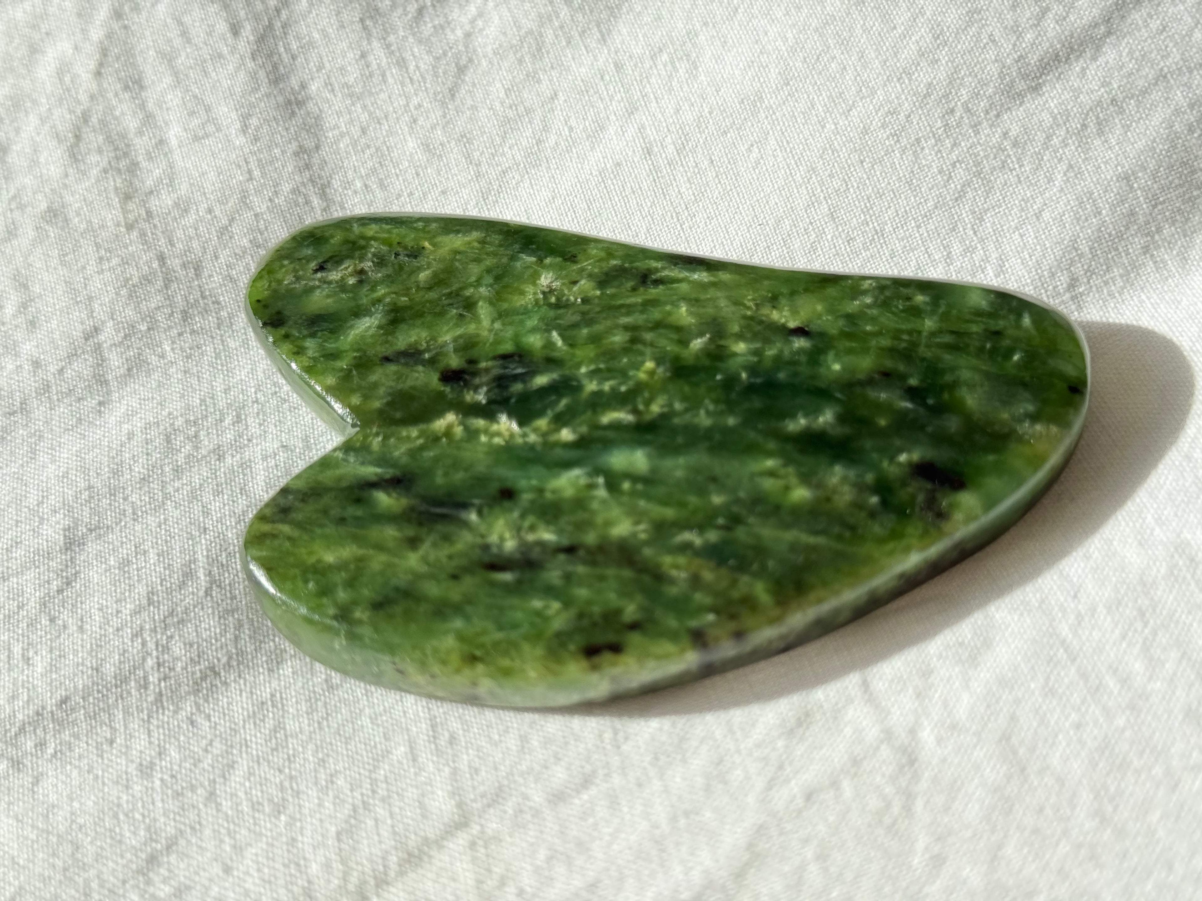 A heart-shaped green jade Gua sha massage tool resting on a textured white linen fabric background.