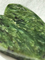A close-up of a polished green stone or jade-like piece with natural marbled texture and dark inclusions, photographed against a light neutral background.