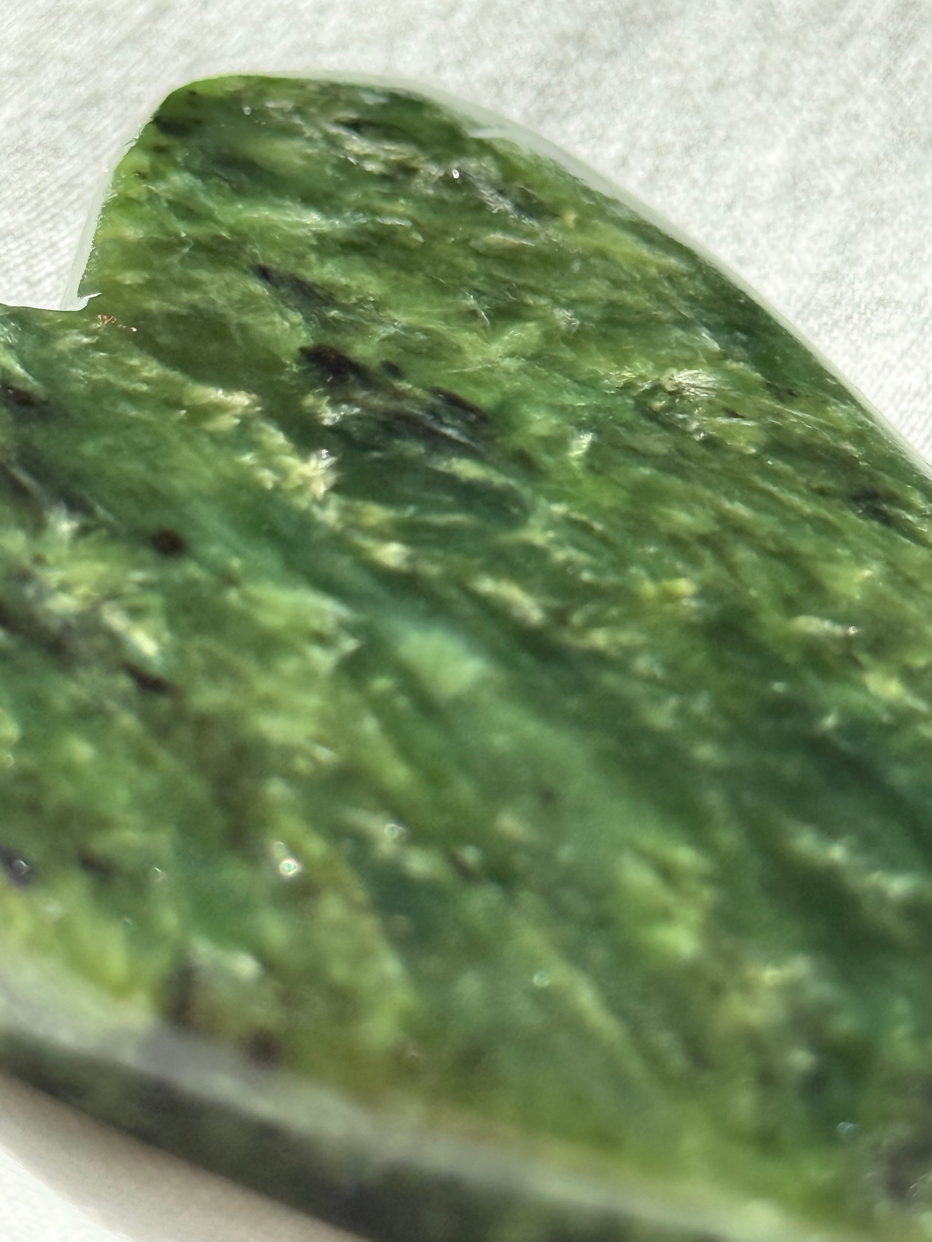 A close-up of a polished green stone or jade-like piece with natural marbled texture and dark inclusions, photographed against a light neutral background.