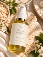A bottle of Tiny Leaf NZ Pure & Sensitive Body Oil with a white pump and clear cap, filled with golden-yellow oil, styled on a textured beige surface with oats, white flowers, and green leaves.