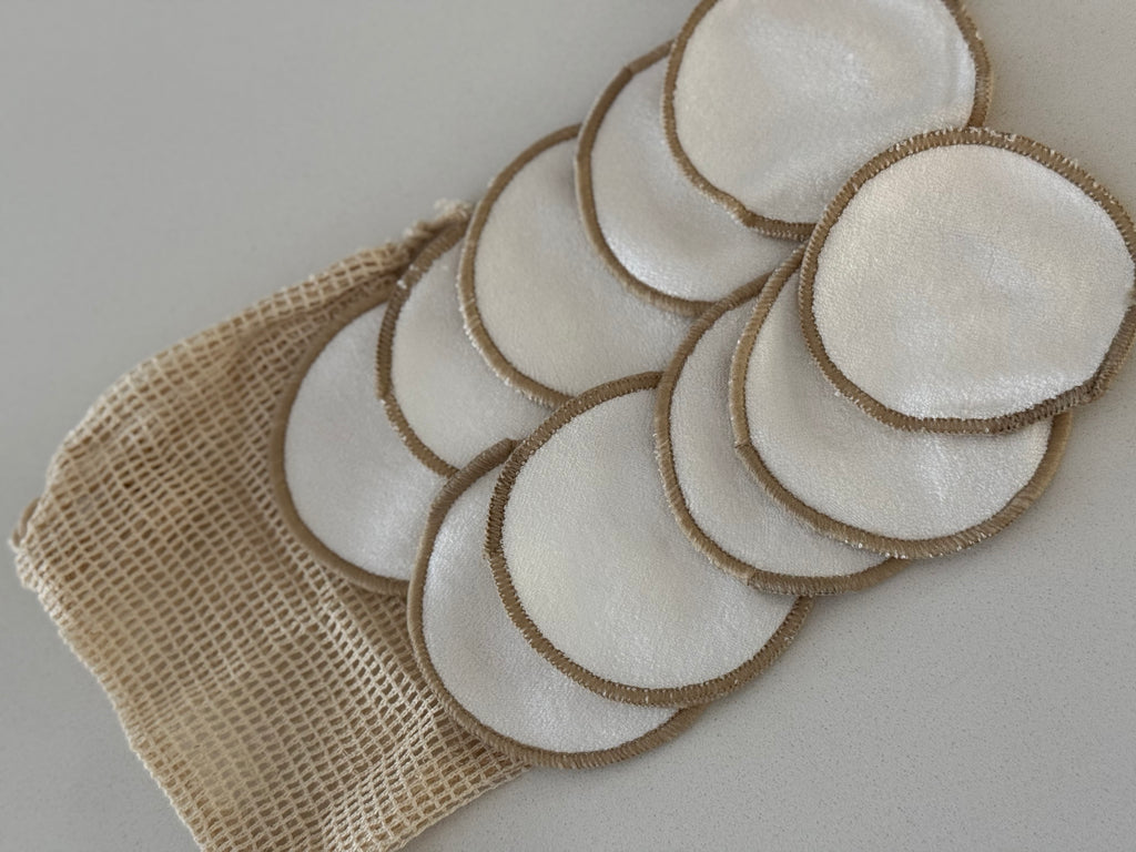 A set of reusable cotton makeup remover pads with natural jute trim, arranged on a beige mesh bag against a light gray background.