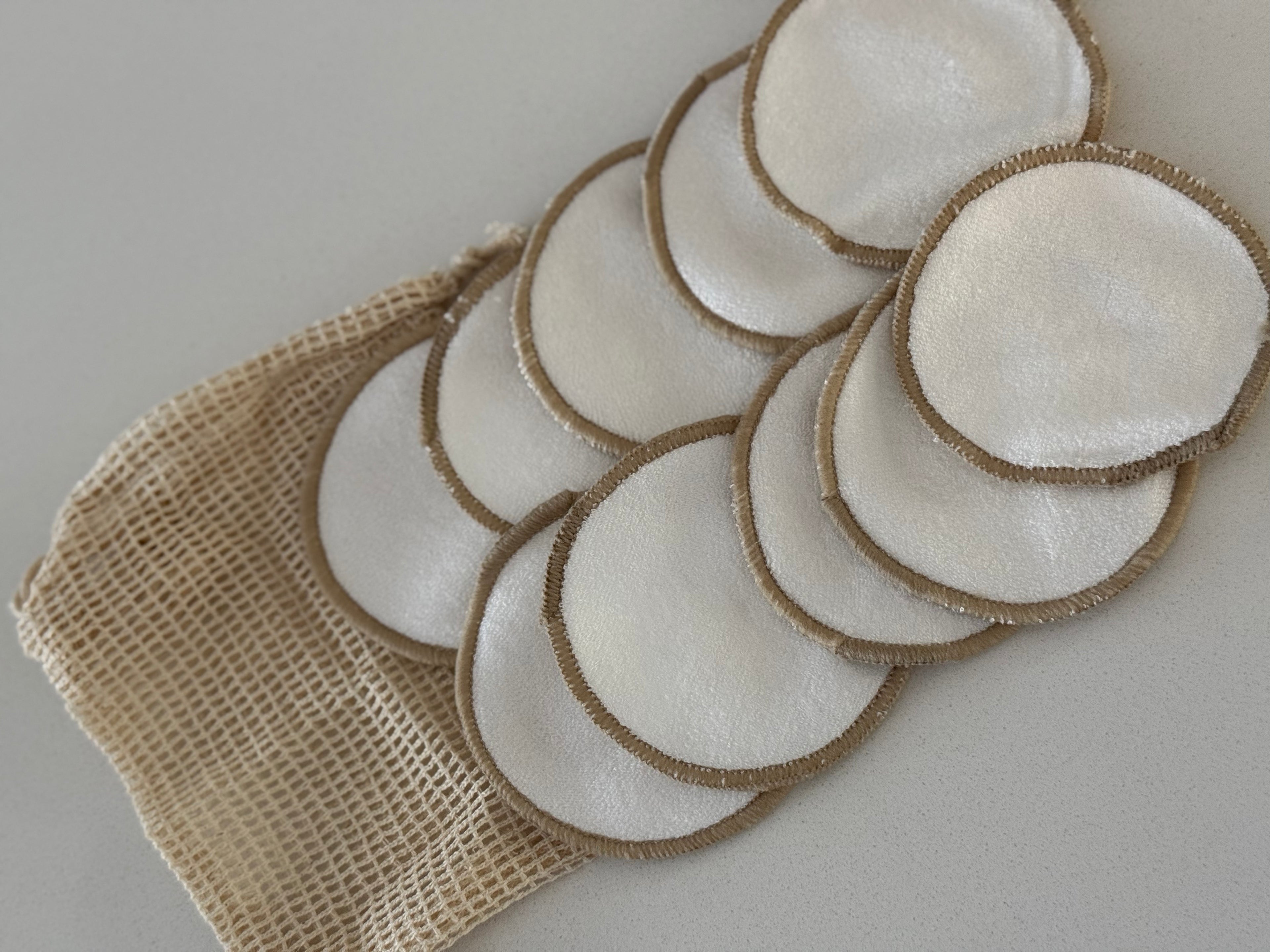 A set of reusable cotton makeup remover pads with natural jute trim, arranged on a beige mesh bag against a light gray background.