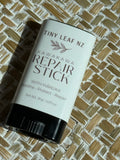 A cylindrical skincare stick container labeled 'Tiny Leaf Nz Kawakawa Repair Stick' with a white label and black cap, placed on a woven straw mat background.