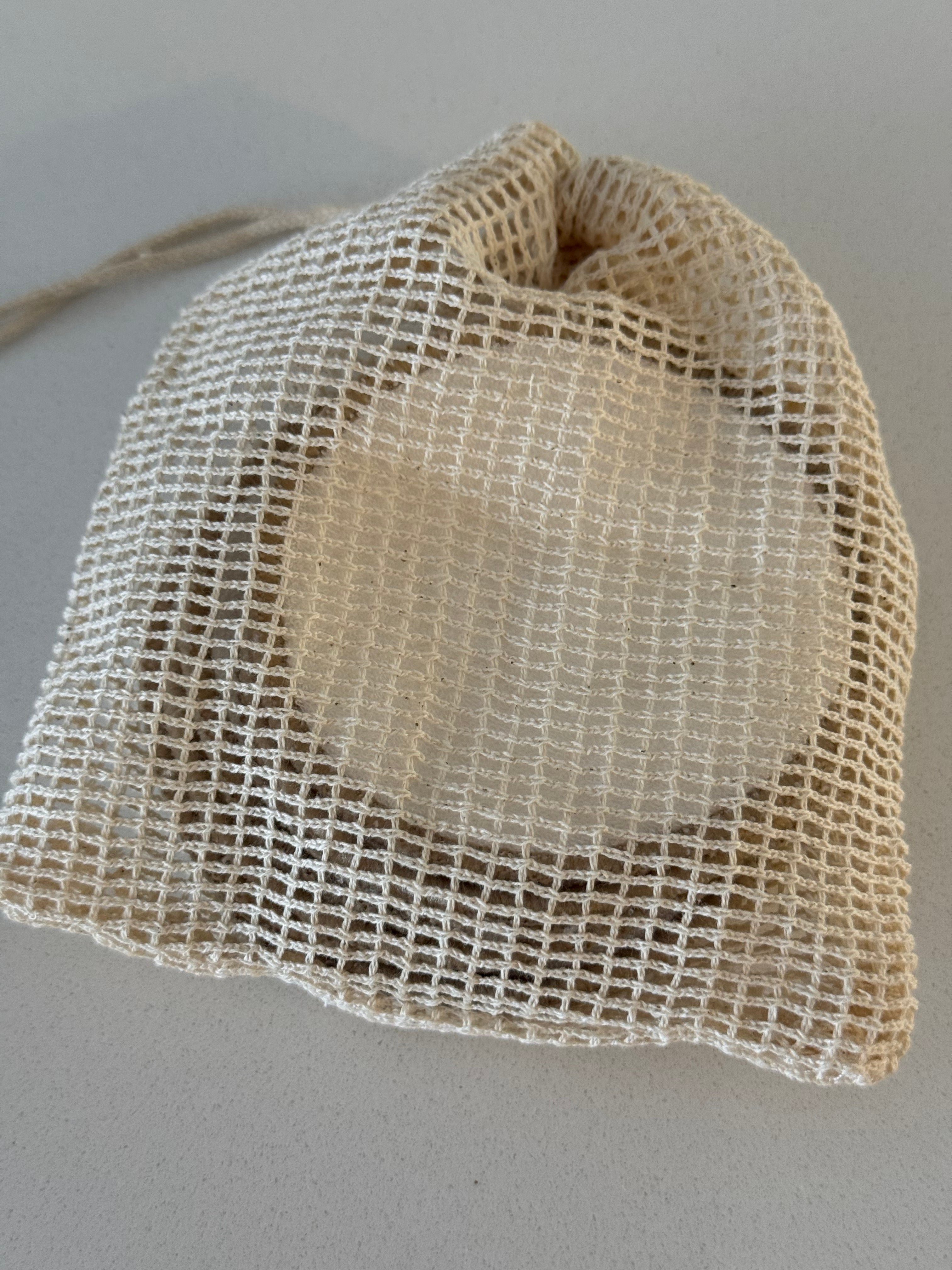 A round, light beige cosmetic sponge or pad enclosed in a small, drawstring beige mesh bag, photographed on a neutral gray background.