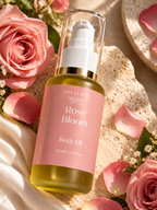 A bottle of Tiny Leafz Rose Bloom Body Oil, featuring a pink label and clear cap, styled with pink roses and petals on a textured beige surface.