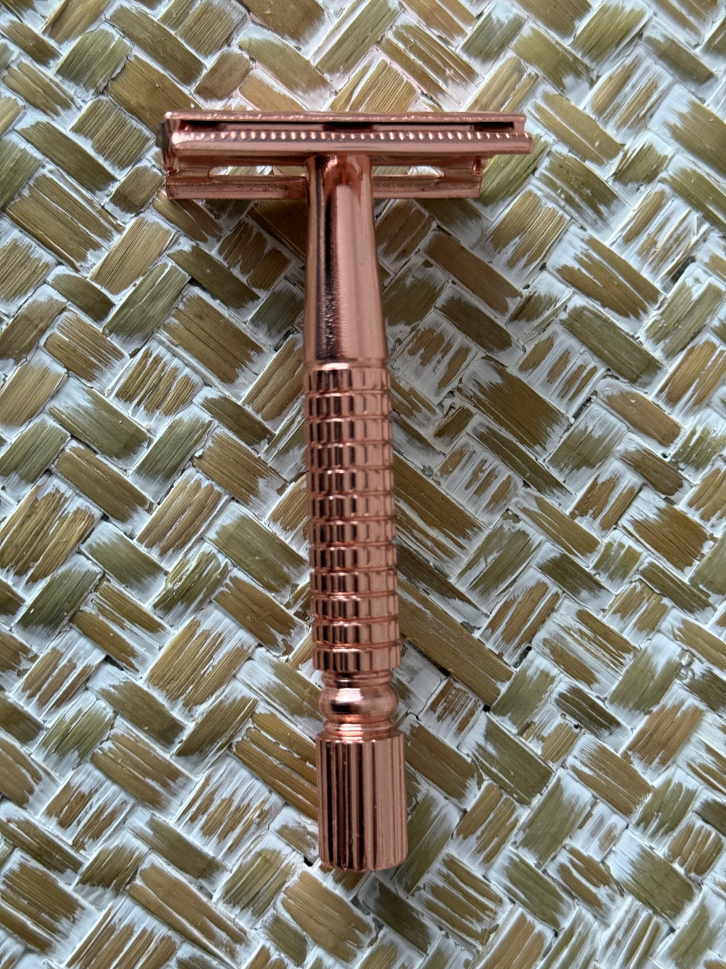A rose gold double-edge safety razor with a ribbed handle, positioned vertically on a woven straw mat background.