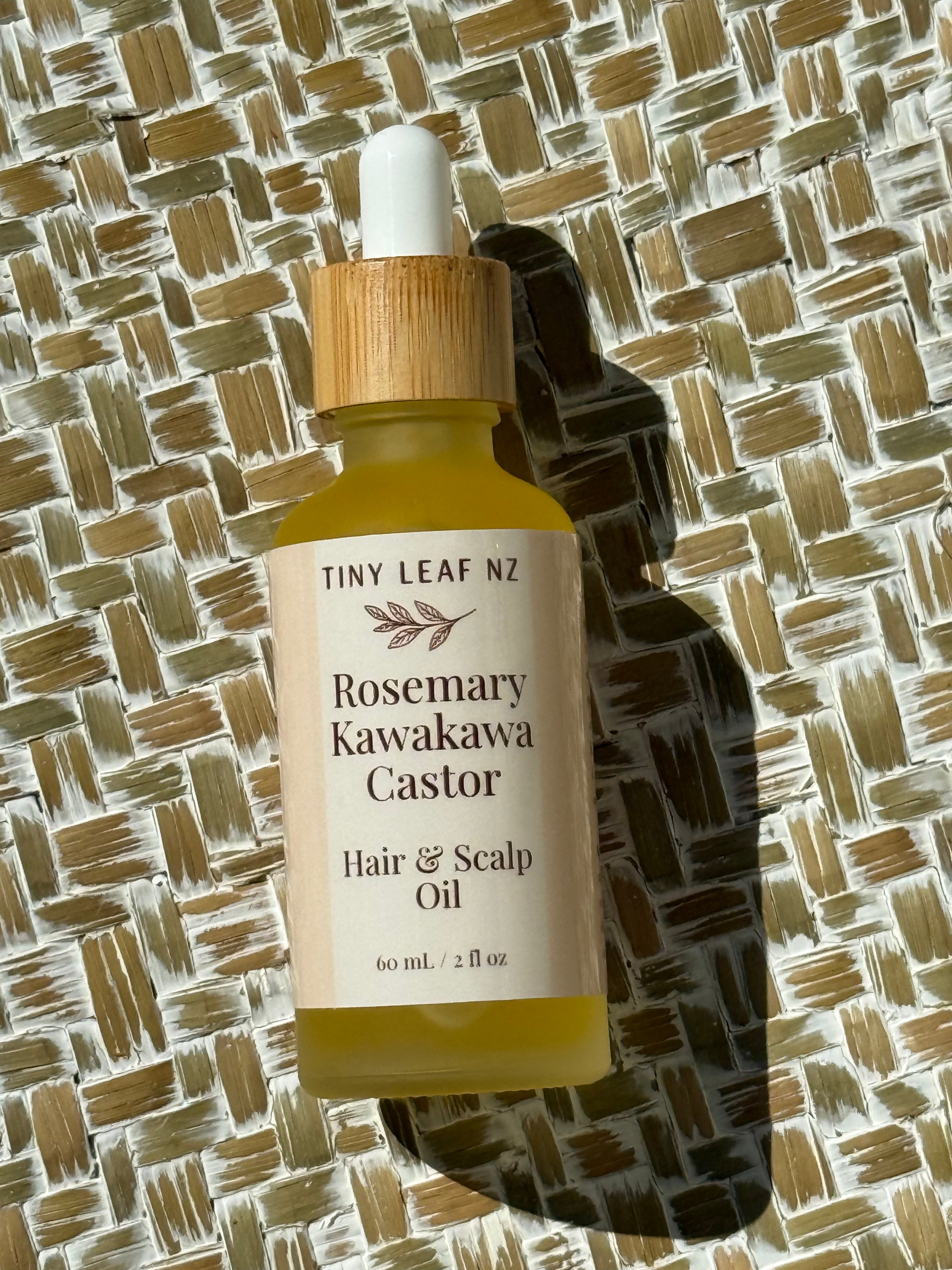 A bottle of Tiny Leaf N2 Rosemary Kawakawa Castor Hair & Scalp Oil, featuring a light wood dropper cap and a cream-colored label, set against a woven straw background with a strong shadow.