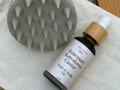 Bottle of hair care product with wooden cap and textured round brush on a cloth surface.