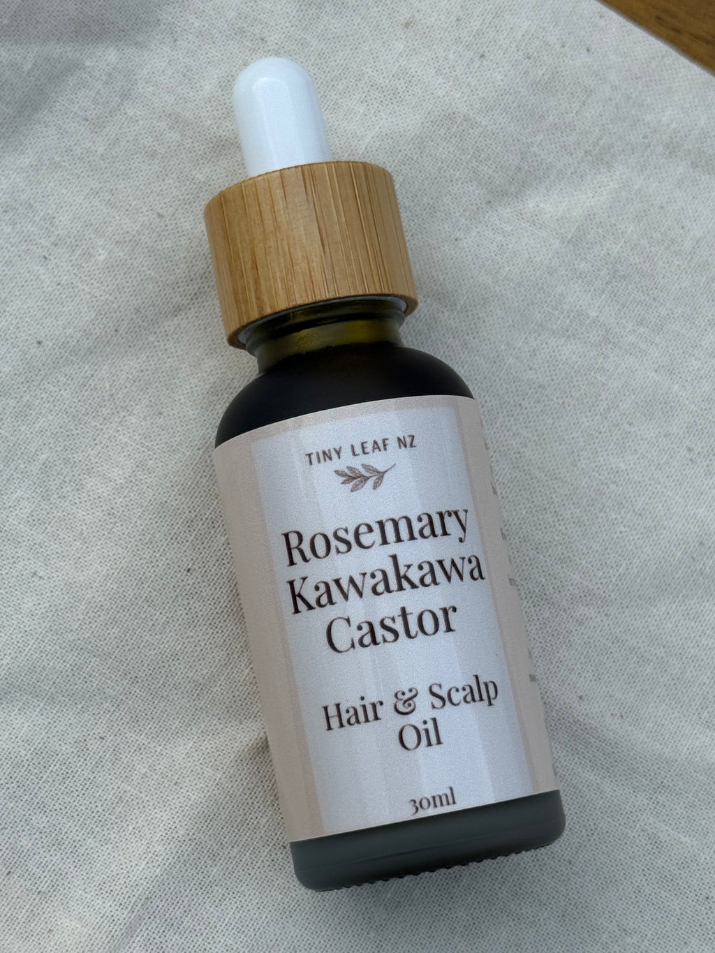 Bottle of Rosemary Kawakawa Castor Hair & Scalp Oil with a dropper on a textured fabric background