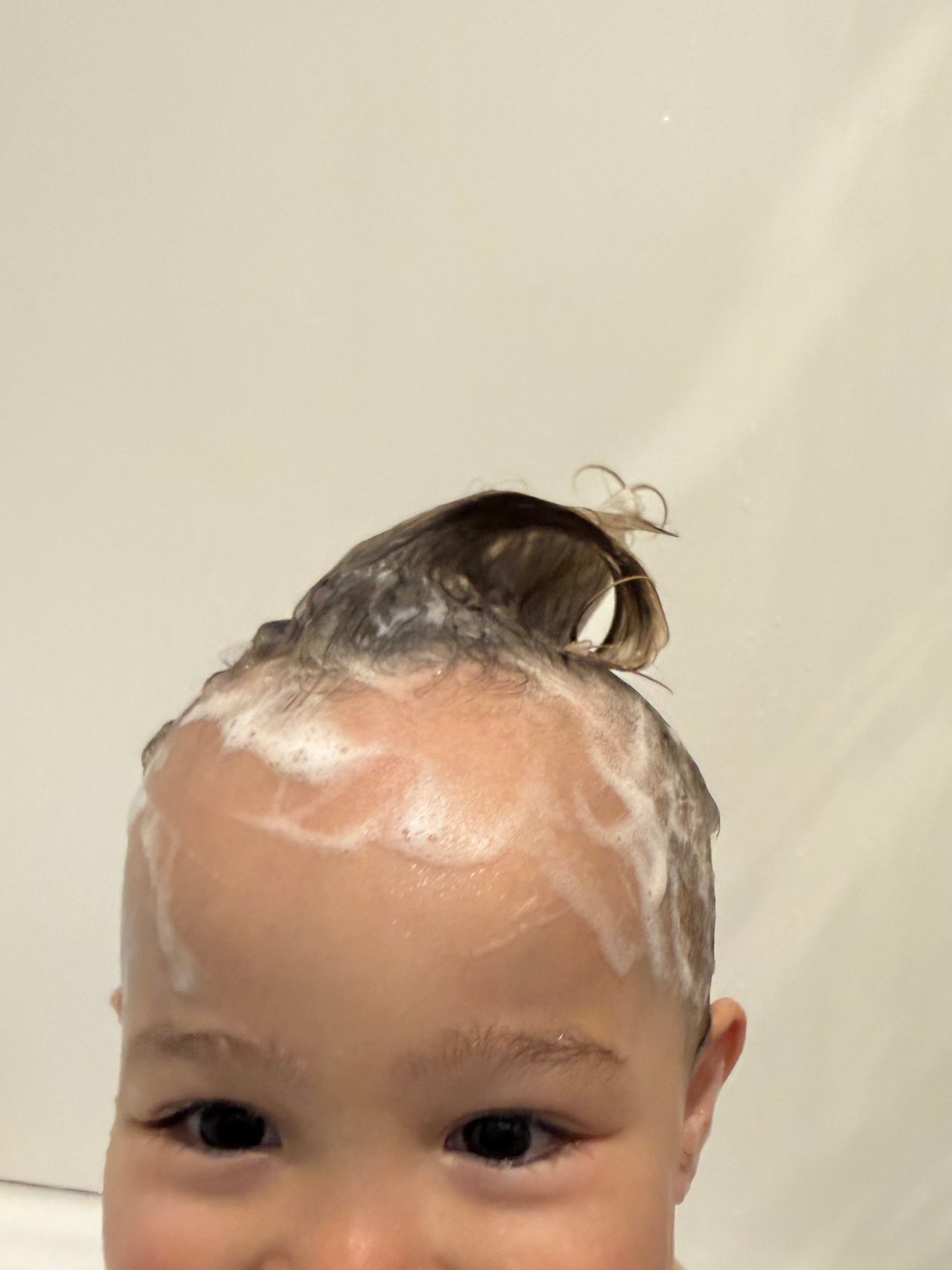 A close-up of a baby with wet hair and white shampoo lather on the scalp, smiling in a bathroom setting.