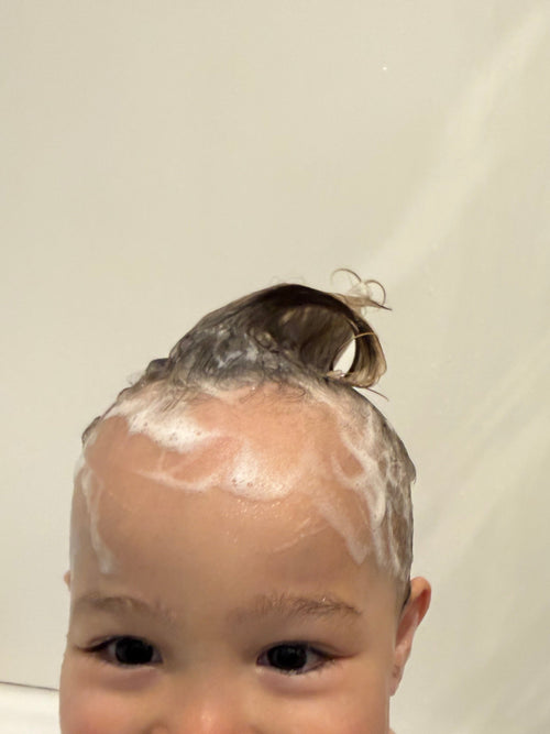 A close-up of a baby with wet hair and white shampoo lather on the scalp, smiling in a bathroom setting.