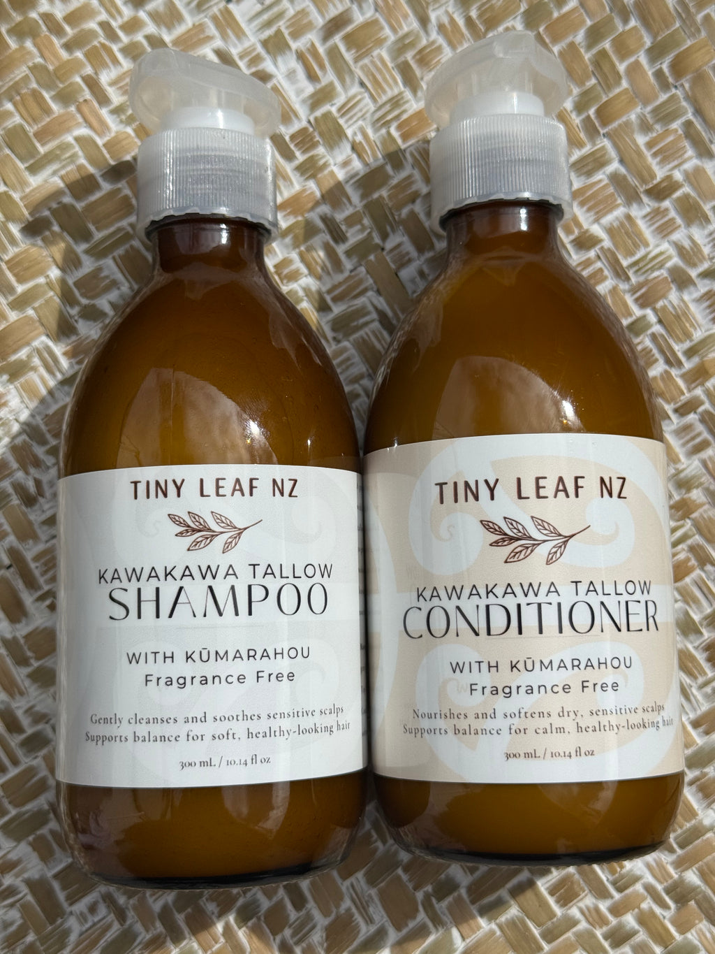 Two amber plastic bottles of Tiny Leaf Nz hair care products, labeled Kawakawa Tallow Shampoo and Kawakawa Tallow Conditioner, placed side by side on a woven tan mat.