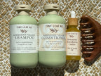 Three Tiny Leaf Nz hair care products arranged on a woven mat: a large bottle of Kawakawa Tallow Shampoo, a large bottle of Kawakawa Tallow Conditioner, and a smaller bottle of Rosemary Kawakawa Castor Hair & Scalp Oil, alongside a dark wooden comb.