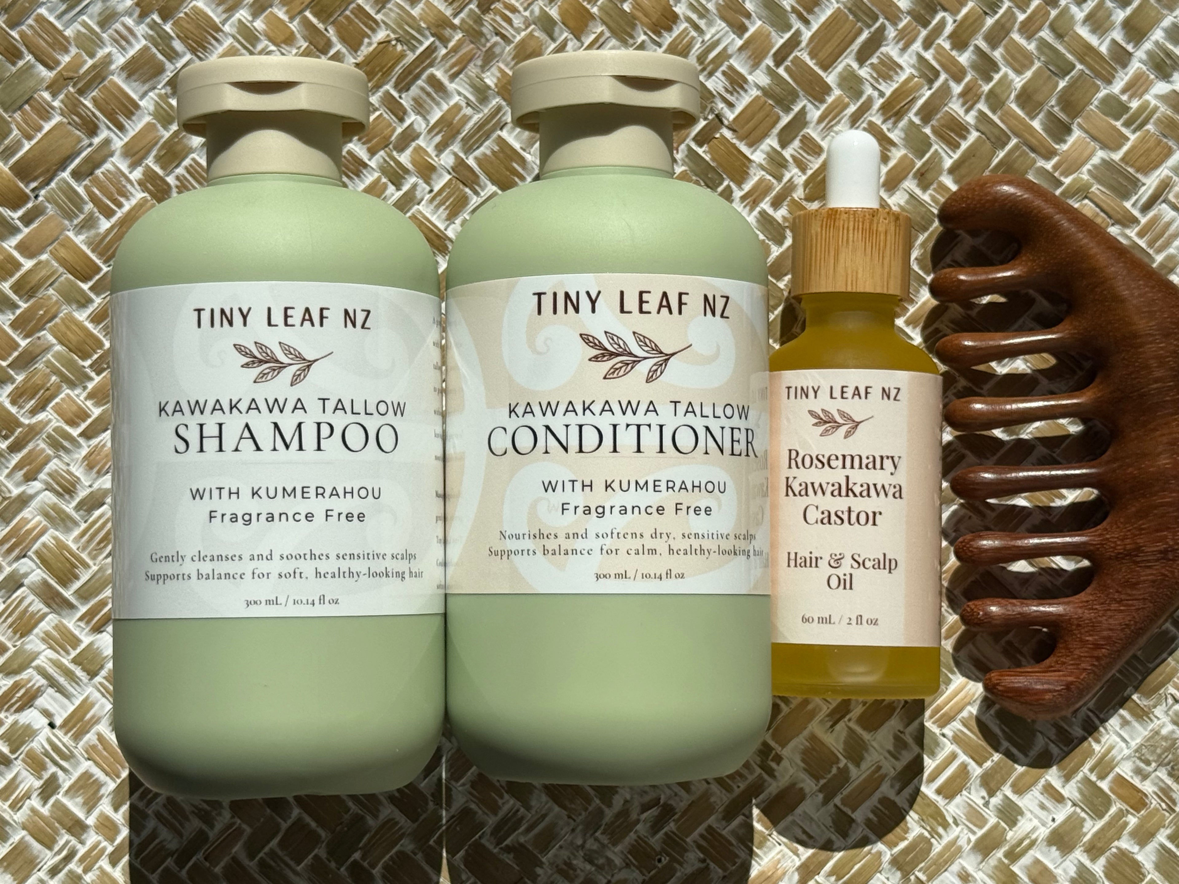 Three Tiny Leaf Nz hair care products arranged on a woven mat: a large bottle of Kawakawa Tallow Shampoo, a large bottle of Kawakawa Tallow Conditioner, and a smaller bottle of Rosemary Kawakawa Castor Hair & Scalp Oil, alongside a dark wooden comb.