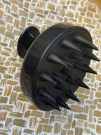 Black silicone scalp massage brush with multiple pointed silicone spikes on a textured beige and white woven mat background.
