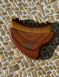 A wooden hair comb with brown bristles and a medium brown wooden handle, featuring the engraved brand name 'TINY LEAF NZ' and a small leaf design, resting on a woven straw mat.