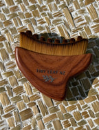 A wooden hair comb with brown bristles and a medium brown wooden handle, featuring the engraved brand name 'TINY LEAF NZ' and a small leaf design, resting on a woven straw mat.