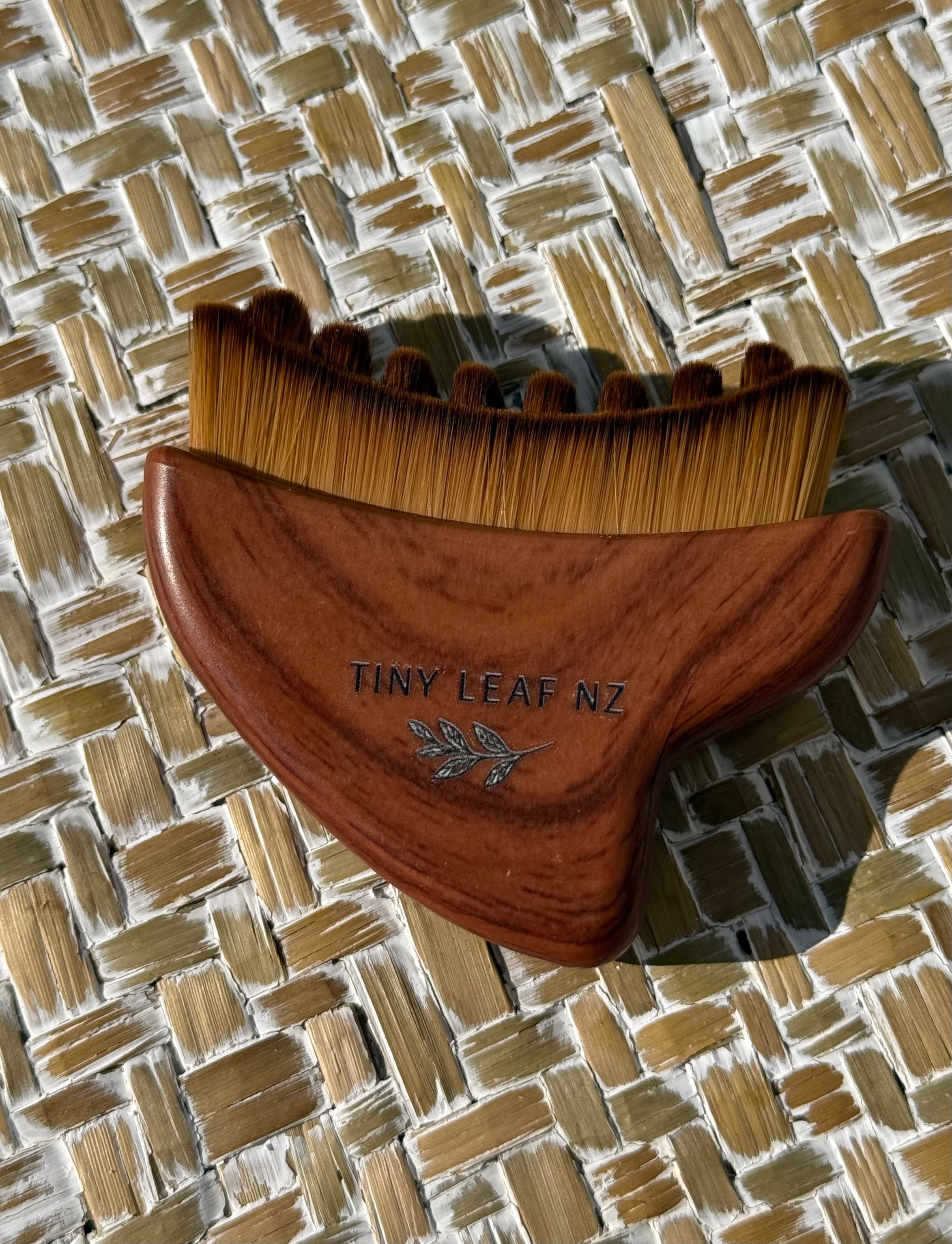 A wooden hair comb with brown bristles and a medium brown wooden handle, featuring the engraved brand name 'TINY LEAF NZ' and a small leaf design, resting on a woven straw mat.