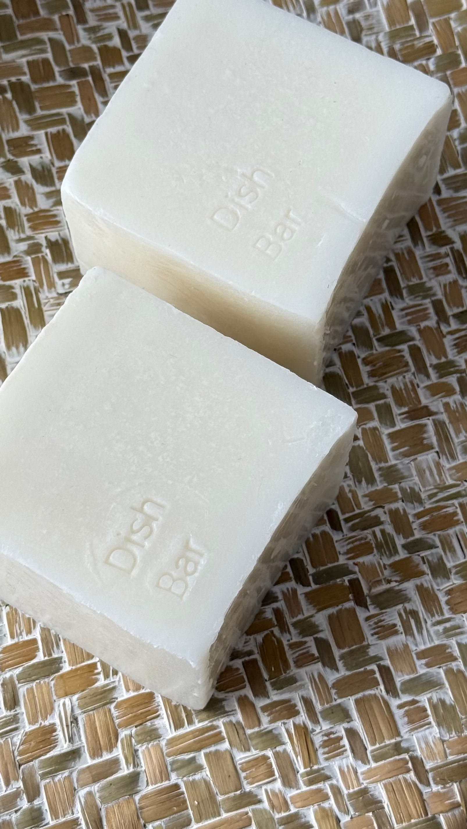 Two rectangular bars of off-white bath bar soap resting on a woven beige and white mat over a wooden surface.