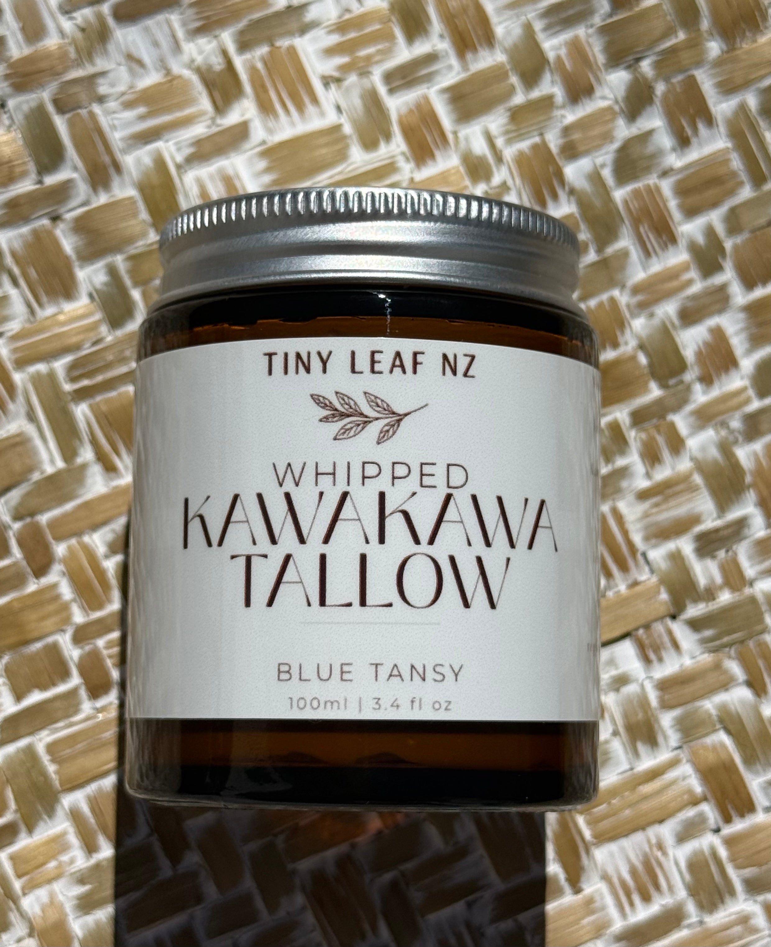 A glass jar candle with a white label reading 'TINY LEAF NZ WHIPPED KAWAKAWA TALLOW BLUE TANSY 100ml | 3.4 fl oz' on a woven straw mat background.