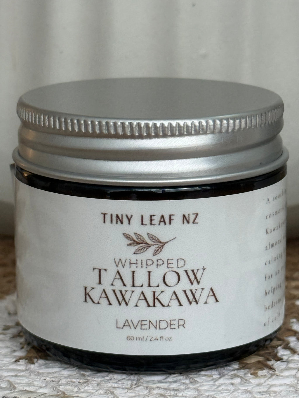 A small dark glass jar with a silver metal lid and a white label reading 'TINY LEAF NZ WHIPPED TALLOW KAWAKAWA LAVENDER' on a textured light surface.