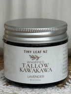 A small dark glass jar with a silver metal lid and a white label reading 'TINY LEAF NZ WHIPPED TALLOW KAWAKAWA LAVENDER' on a textured light surface.