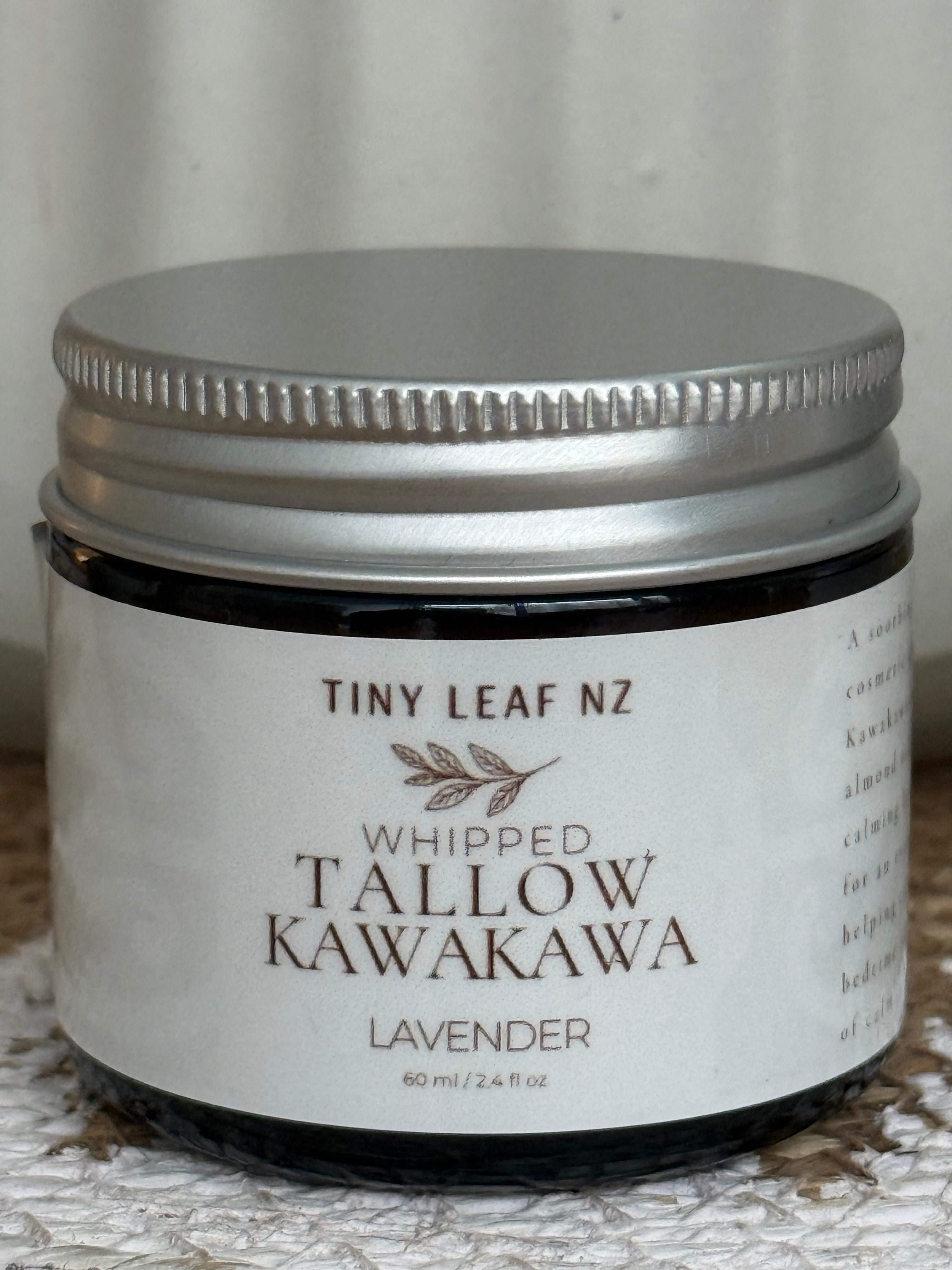 A small dark glass jar with a silver metal lid and a white label reading 'TINY LEAF NZ WHIPPED TALLOW KAWAKAWA LAVENDER' on a textured light surface.