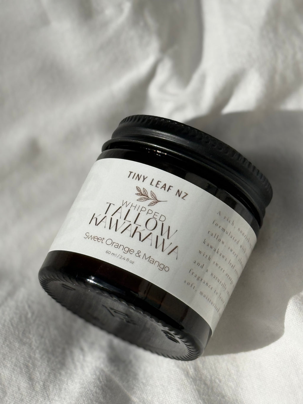 A jar of Tiny Leaf NZ Whipped Tallow Manuka Sweet Orange & Mango scented soap, shown on a textured white fabric background with a soft shadow.
